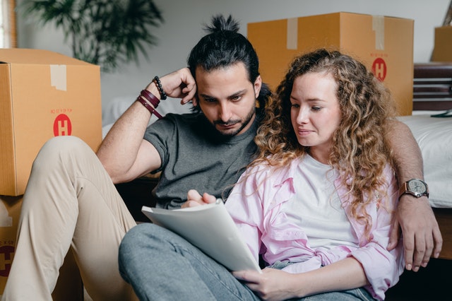 A couple sitting beside packed boxes and writing in a notebook the perks of a condo or a house.