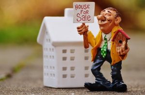 A toy figurine of a person holding a “House for sale” sign next to a white toy house.